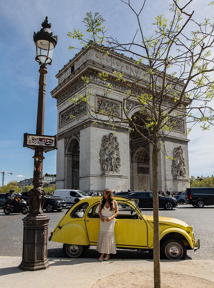 Guest standing beside a vintage yellow Citroën 2CV in front of the Arc de Triomphe on a sunny day in Paris.
