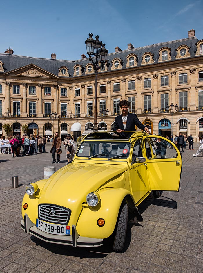 Person standing through the open roof of a vintage yellow Citroën 2CV in Place Vendôme, with elegant Parisian architecture and pedestrians in the background.