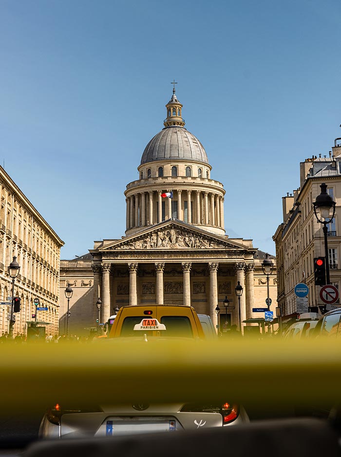 The Panthéon in Paris viewed from the street with a yellow Paris taxi in the foreground and its neoclassical dome rising against a clear blue sky.