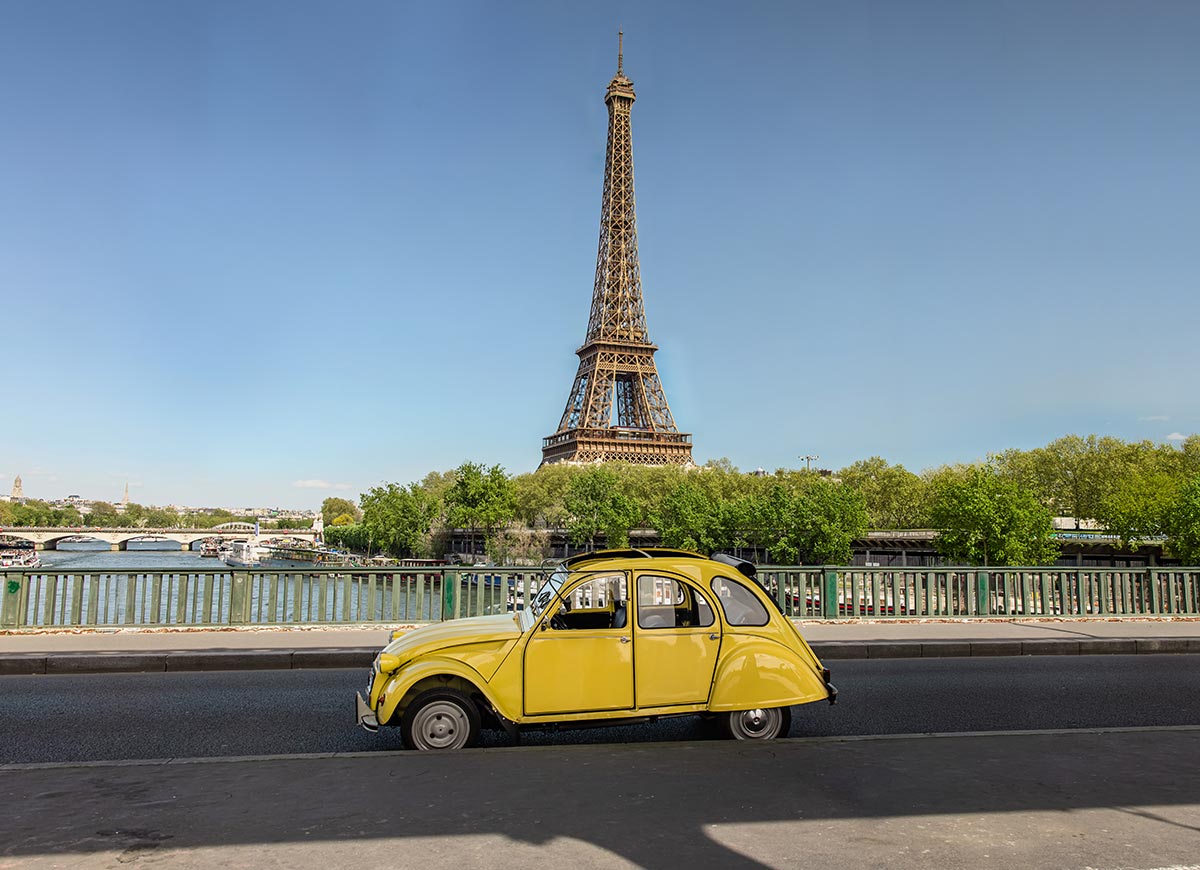 Vintage yellow Citroën 2CV parked beside the Seine River with the Eiffel Tower rising above the trees under a clear blue Paris sky.
