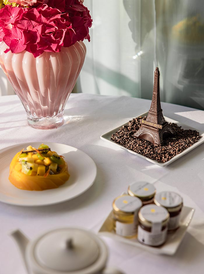 Close up of a breakfast table featuring a chocolate Eiffel Tower sculpture on a bed of cocoa nibs, a fresh fruit tart, and small jars of preserves. Bright pink flowers in a glass vase and soft morning sunlight create an elegant Parisian dining setting.