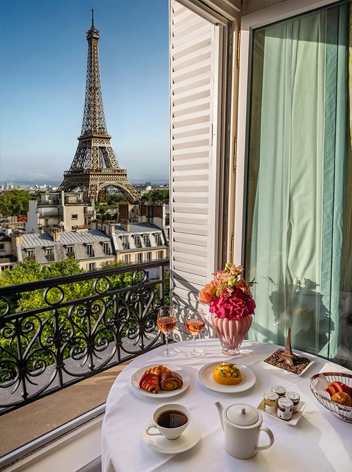 Breakfast table set with coffee, pastries, flowers, and champagne beside an open balcony overlooking the Eiffel Tower. The iconic Paris landmark rises above surrounding rooftops under a clear blue sky, creating a classic Paris hotel view.