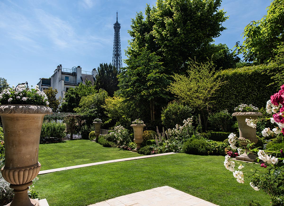 Manicured garden with lush green lawns, flowering shrubs, and decorative stone planters beneath a clear blue sky. The Eiffel Tower rises above the trees in the background, creating a peaceful Parisian garden scene.