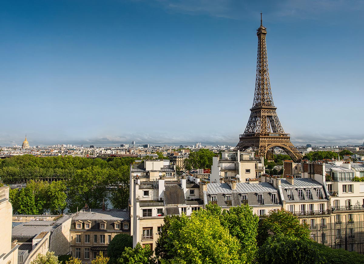The Eiffel Tower rises above Paris rooftops and tree lined streets on a clear day, with the city skyline stretching into the distance beneath a blue sky. Sunlight highlights the iron lattice structure while classic Parisian buildings fill the foreground.