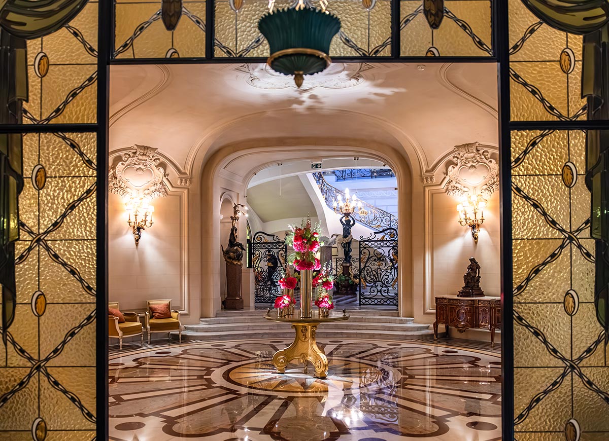 Grand hotel lobby with polished marble floors, ornate wall sconces, and an elegant floral arrangement displayed on a central table. Decorative ironwork railings, sculptural details, and stained glass panels create a luxurious Parisian atmosphere.