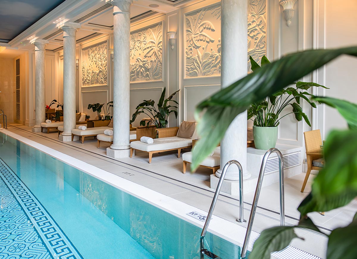 Elegant indoor swimming pool lined with white marble columns, cushioned lounge chairs, and tropical plants. Decorative wall panels and soft lighting create a luxurious spa atmosphere beside the turquoise water.