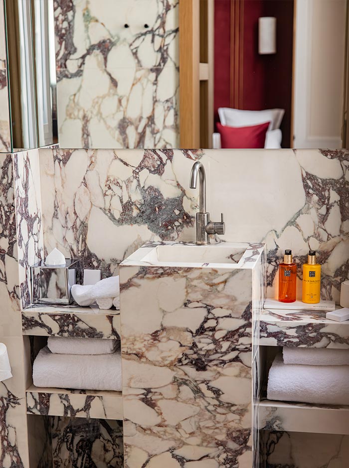 Luxury marble bathroom vanity with dramatic veined stone surfaces, a modern square sink, and neatly arranged towels and bath amenities. A large mirror reflects warm interior details, creating an elegant and sophisticated hotel bathroom setting.