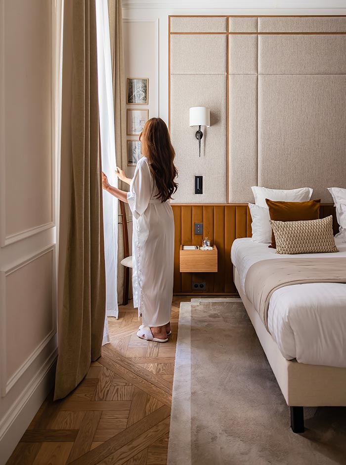 Person in a white robe standing beside a large window in a bright hotel bedroom, looking out through floor to ceiling curtains. The room features a plush bed with layered pillows, warm wood accents, and elegant neutral decor that creates a calm and luxurious atmosphere.