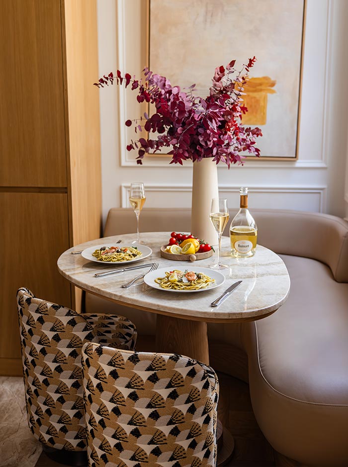 Elegant dining nook with a marble table set for two, featuring plates of pasta, champagne, and fresh fruit. A large floral arrangement in deep pink tones serves as the centerpiece, while curved banquette seating and contemporary artwork create a refined and intimate atmosphere.