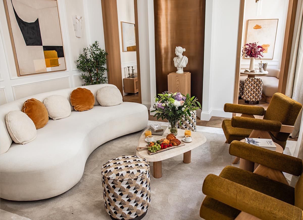 Elegant hotel suite living room with a curved cream sofa, velvet armchairs, and contemporary artwork in a bright, refined setting. A coffee table topped with pastries, fruit, flowers, and coffee creates a welcoming atmosphere, while sculptural decor and warm wood accents add a luxurious touch.