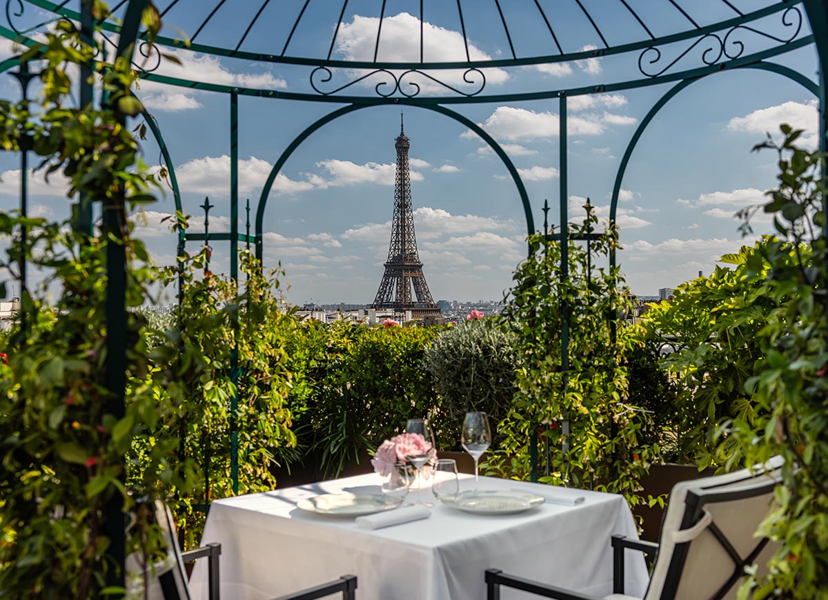 Romantic rooftop dining table set for two beneath a wrought iron gazebo, with the Eiffel Tower perfectly framed in the distance. Lush greenery surrounds the terrace while a white tablecloth, place settings, and pink flowers create an intimate Parisian dining scene.