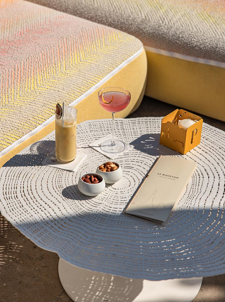 White rooftop lounge table set with a pink cocktail, iced coffee, mixed nuts, and a menu labeled "LE ROOFTOP." Curved yellow cushioned seating and warm afternoon sunlight create an upscale outdoor terrace atmosphere.