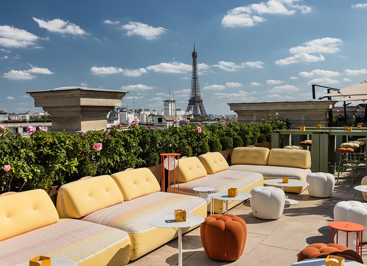 Elegant rooftop terrace with curved yellow lounge seating, white tables, and flowering greenery overlooking the Eiffel Tower under a bright blue sky with scattered clouds. The open air Paris rooftop setting offers panoramic city views and a relaxed luxury atmosphere.