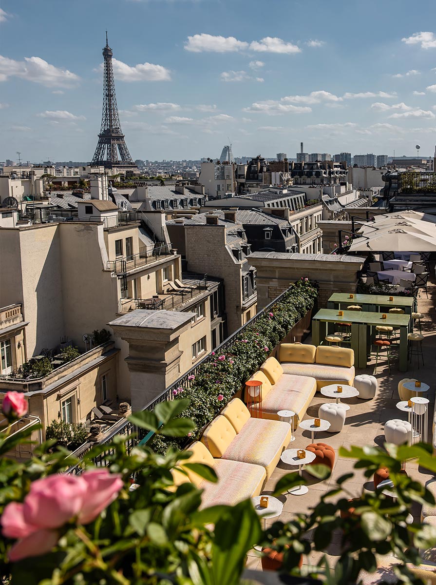 Rooftop terrace overlooking Paris with the Eiffel Tower rising above the city skyline in the distance. Soft yellow lounge seating, dining tables, and pink roses create an elegant outdoor space with panoramic views of the surrounding rooftops.
