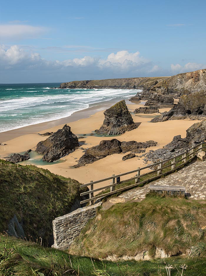 Golden sandy beach dotted with dramatic dark rock formations beneath rugged coastal cliffs as turquoise waves roll onto the shore. A stone path and wooden fence overlook the sweeping coastline under a bright blue sky with scattered clouds.